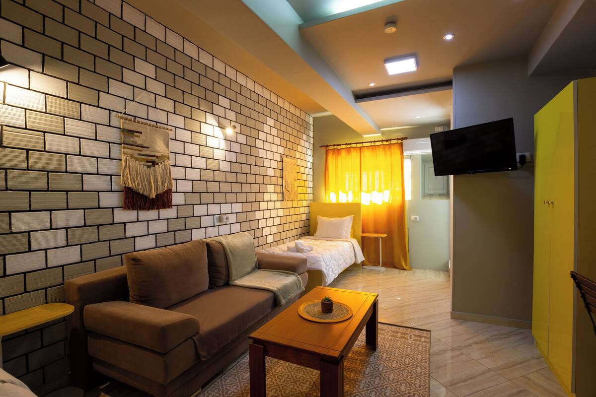 A modern hotel room with a brick wall, a sofa, a bed, a TV, and a small table with a plant.