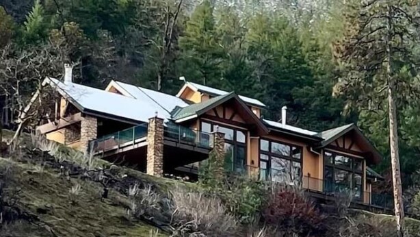A modern cabin with a stone foundation, large windows, and a sloped roof, surrounded by dense forest.