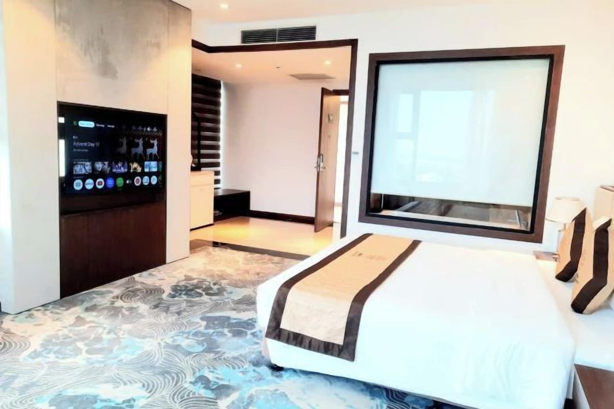 A modern hotel room with a large bed, a flat-screen TV mounted on the wall, and a large window.