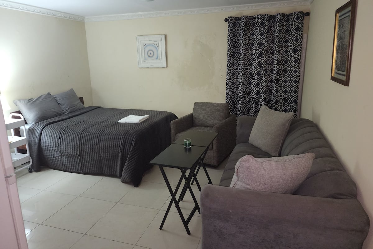 A small, well-lit room with a bed, a sofa, a small table, and a patterned curtain.