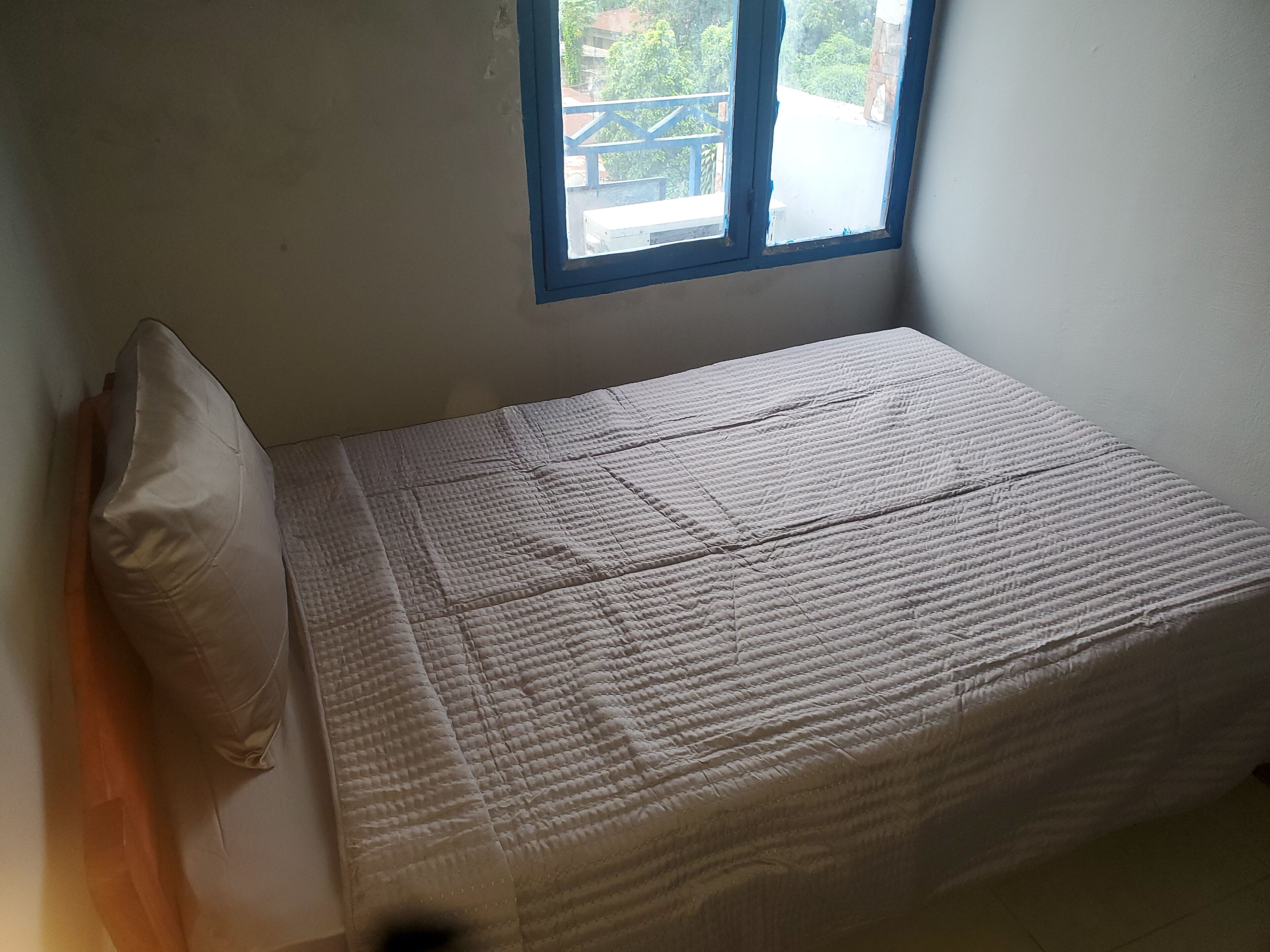Une petite chambre avec un lit simple, un matelas blanc uni et un oreiller. Une fenêtre à cadre bleu est visible, donnant sur un balcon extérieur.