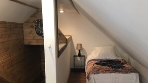 Un dormitori amb sostre inclinat, parets de fusta, un llit amb llençol estampat, una taula de nit amb una làmpada i un rellotge de paret.