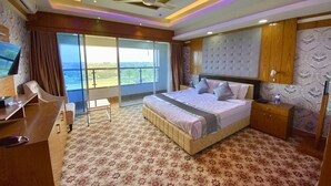 A spacious bedroom with a large bed, a desk, and a chair, a balcony with a view, and modern ceiling lighting.