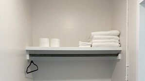 A small, white-walled bathroom with a wooden floor, a white shelf holding towels and toilet paper, and a black hanger on the wall. - Innovative Studio Apartment (Gadsden)