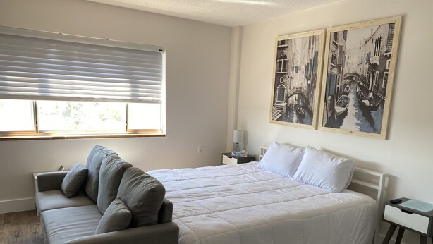 A bedroom with a bed, a sofa, a nightstand, and a window with blinds. - Innovative Studio Apartment (Gadsden)
