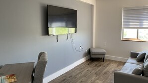 A modern living room with a flat-screen TV mounted on the wall, grey sofas, a wooden floor, and a window with blinds. - Innovative Studio Apartment (Gadsden)