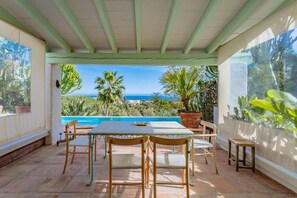 Pátio externo com mesa e cadeiras, com vista para uma piscina e paisagem panorâmica do oceano e colinas.