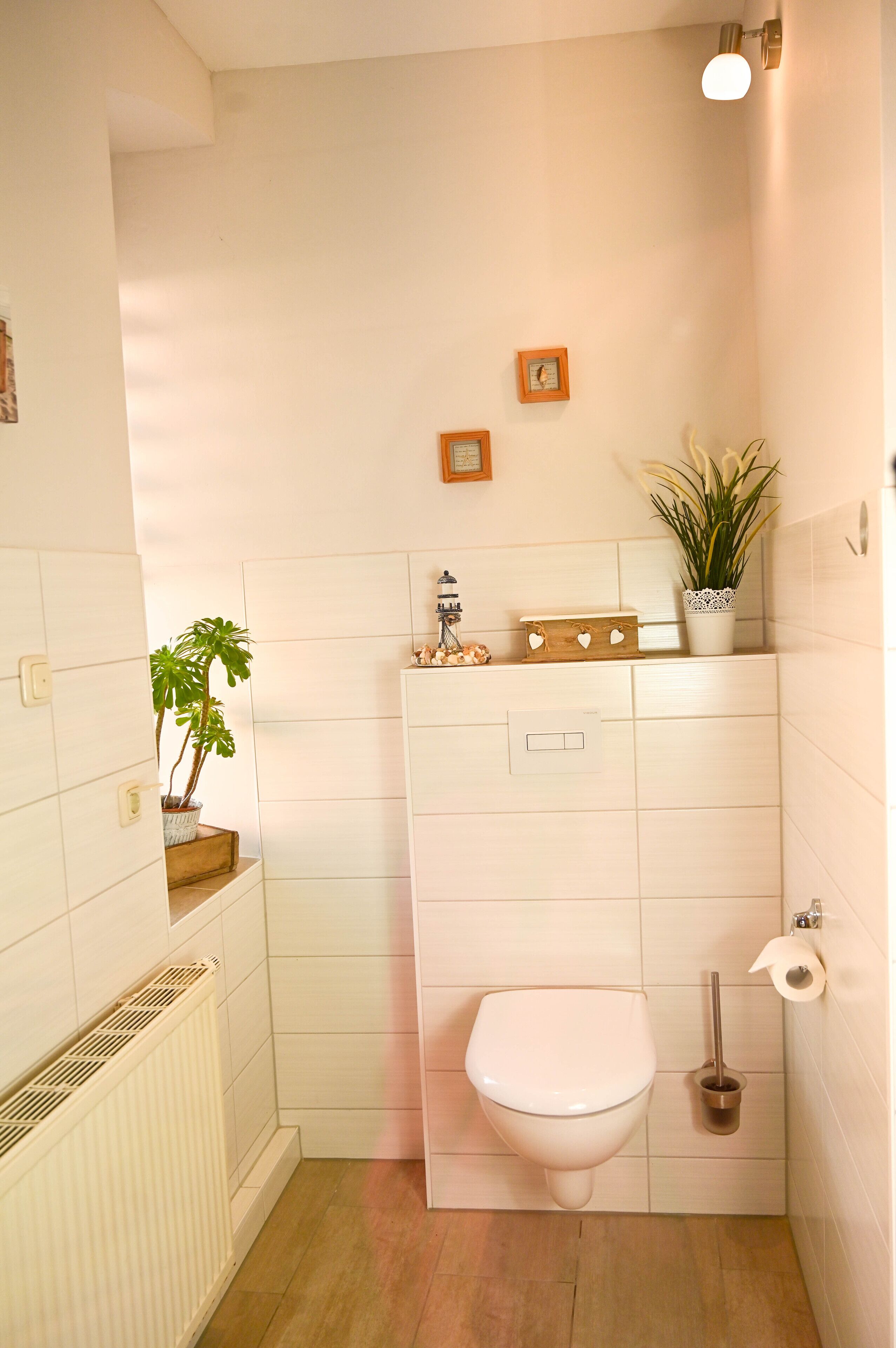 Un baño pequeño y limpio con azulejos blancos, inodoro, radiador, planta en maceta y estante con adornos.