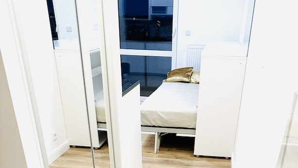 Ein schmaler Flur mit Holzboden führt zu einem Schlafzimmer mit Bett und Fenster.