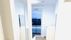 Ein schmales Entree mit Holzboden, weißen Wänden und einem großen Spiegel an einer Seite. Eine Tür führt zu einem Balkon mit Blick auf die Stadtlandschaft.