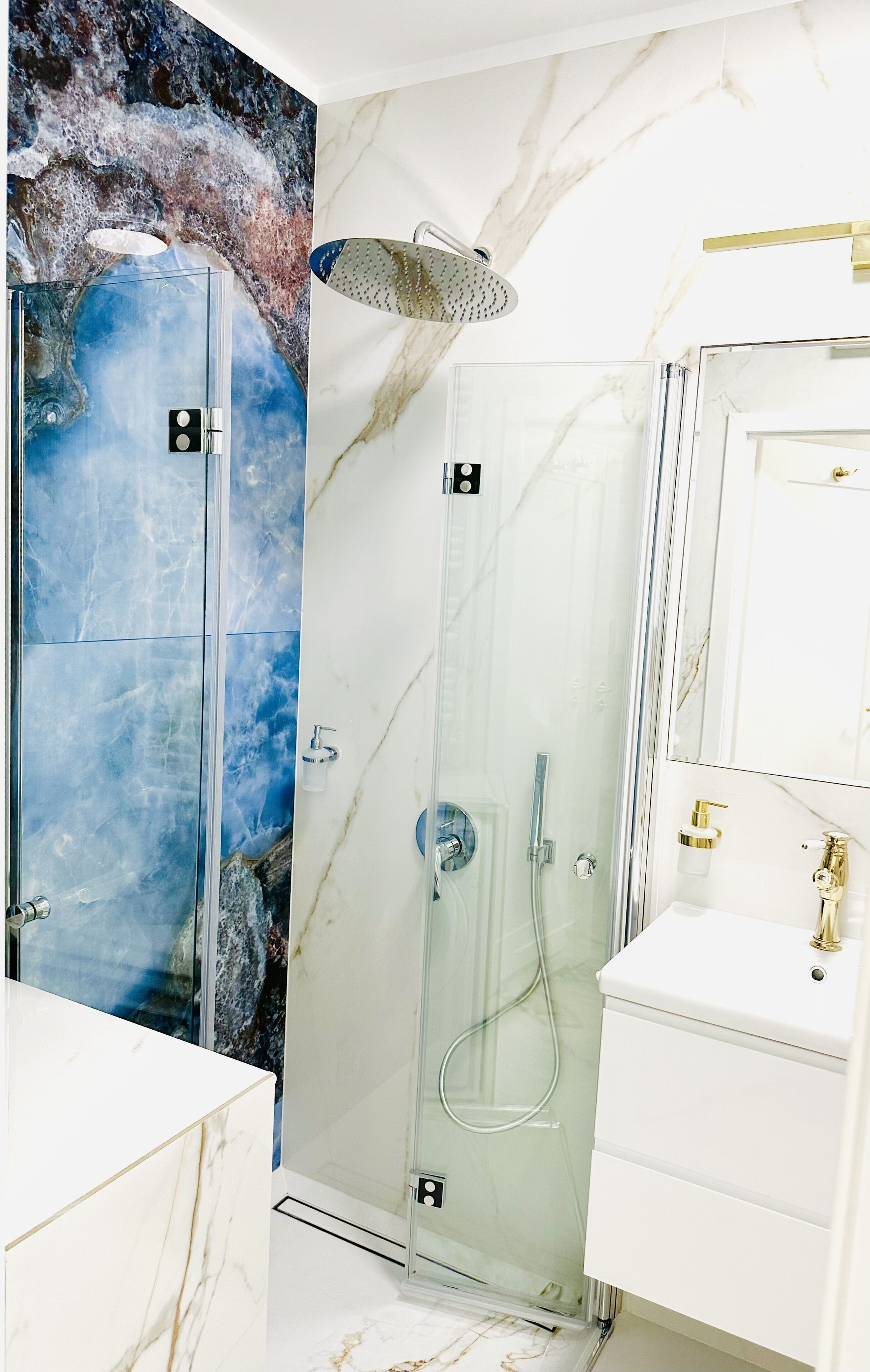 A modern bathroom with a glass-enclosed shower, a marble wall, and a white vanity with a gold faucet.
