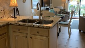 A kitchen with granite countertops, wooden cabinets, a double sink, and a dining area with a table and chairs. - Gorgeous 203C Gulf Shores Surf and Racket Club (Gulf Shores)