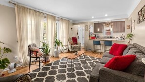 Un salon avec un canapé, une table basse et un tapis à motifs. Au fond, on aperçoit un coin cuisine avec un bar et des chaises.