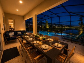 A dining area with a table set for four, a sofa, and a view of a swimming pool at night.