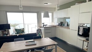A modern kitchen with white cabinets, a wooden dining table, and two chairs. There is a large window providing a view of the outdoors. - La P'Tite Case - facing the sea (Les Sables-d´Olonne)