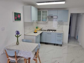 Eine kompakte Küche mit Gaskochfeld, Spüle und hellblauen Schränken. Ein Esstisch mit Stühlen und eine Blumenvase sind ebenfalls vorhanden.