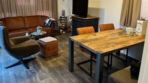 A living room with a brown leather sofa, a wooden coffee table, a dining table with four chairs, a television on a stand, and a bookshelf. - Chalet in the family paradise "Olmenveld", Zeeland (Serooskerke)