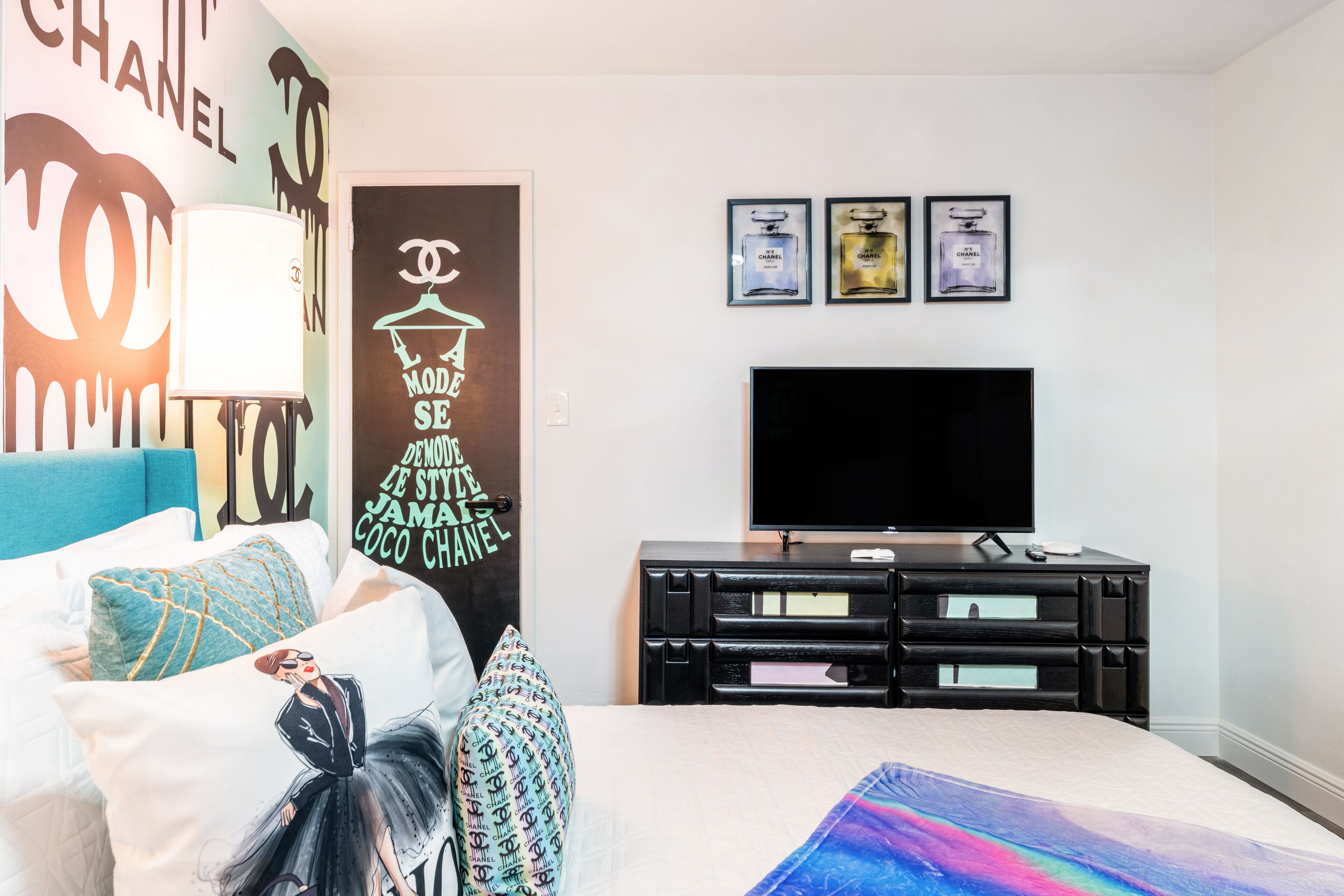 A bedroom with a bed, pillows, a TV, a dresser, and wall art including Chanel branding.