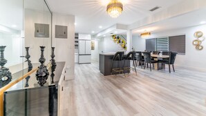 A modern kitchen with a central island, a dining area with a chandelier, and a staircase with decorative elements. - Blackrock Lux ~ High-End Designer Oasis ~ 7min from Airport ~ EV Charger (Fort Lauderdale)