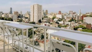 Terraza en la azotea con mesas y sillas de metal blanco, que ofrece una vista panorámica de un paisaje urbano con varios edificios.