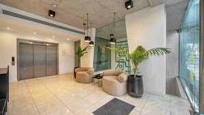 Un vestíbulo moderno con pared de vidrio, una recepción, dos sofás, una mesa de centro, una planta grande en maceta y un ascensor.