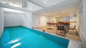 Una piscina interior con una zona soleada que cuenta con una mesa, sillas, una planta pequeña y un suelo de baldosas.