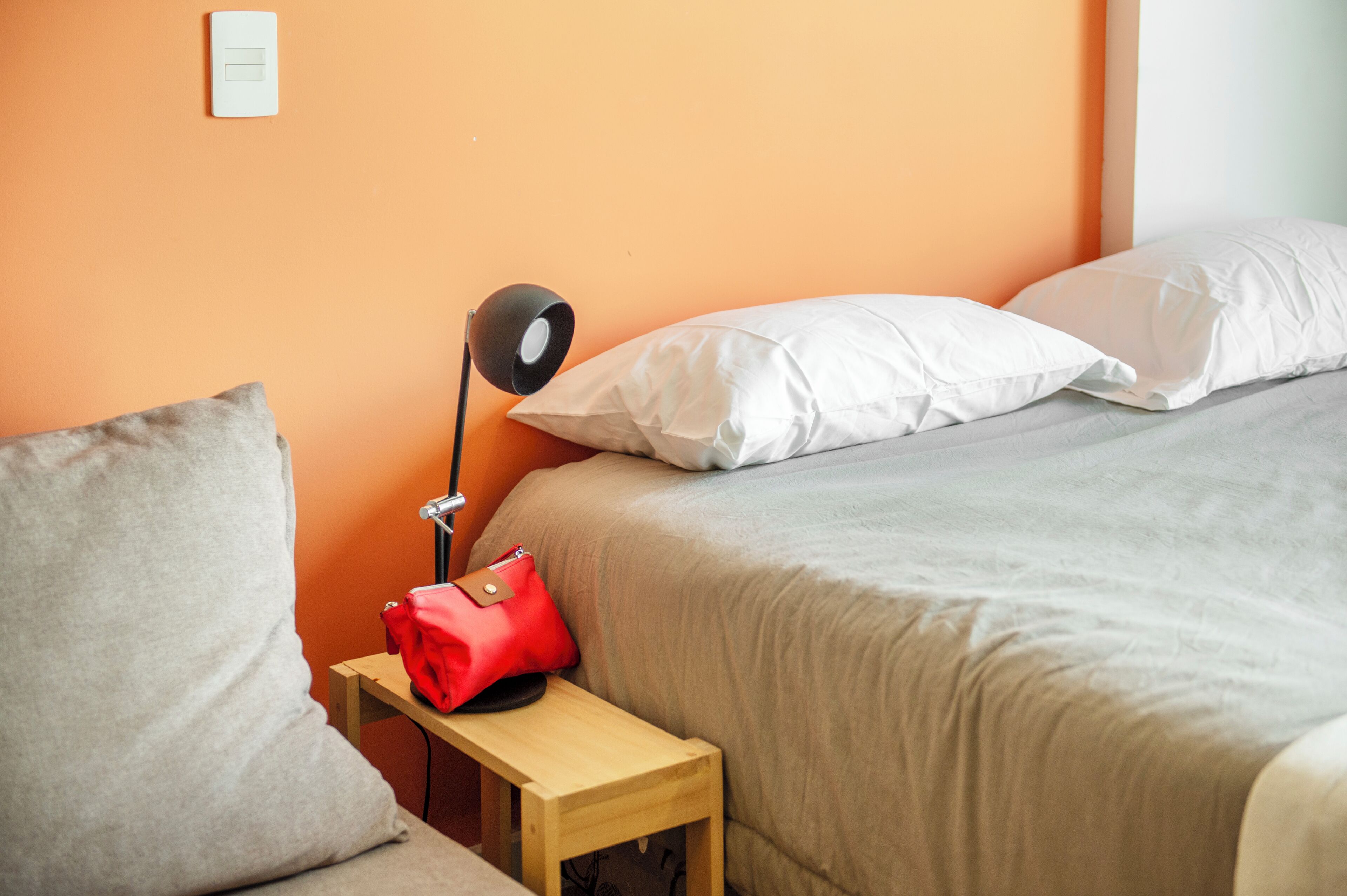 A neatly made bed with white pillows and a beige blanket, a red bag on a small wooden nightstand, and a black desk lamp.