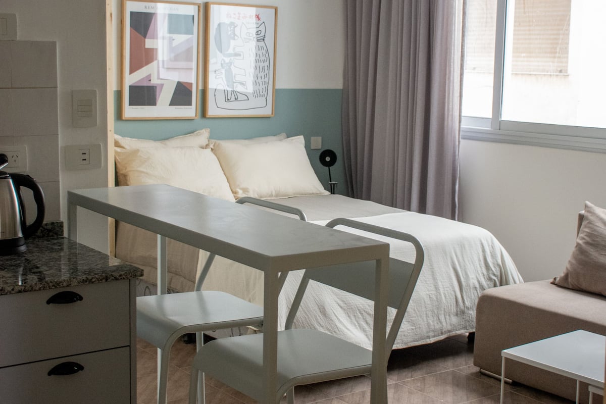A modern, well-lit bedroom with a bed, a desk, and a chair. There is a window with curtains, a radiator, and artwork on the wall.