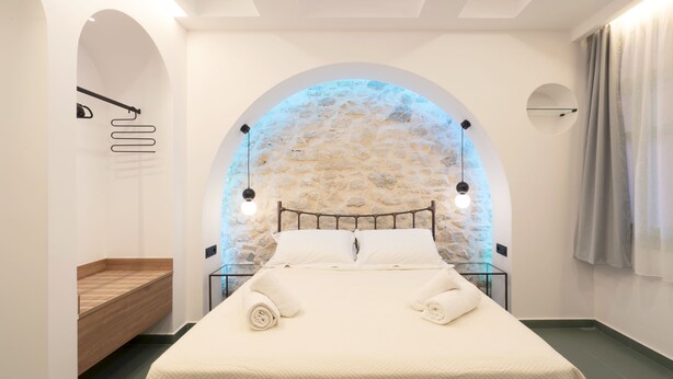 A modern bedroom with a stone wall arch, a bed with white linens, and a wooden bench. - SARTORI CONCEPT HOTEL (Heraklion)