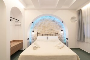 A modern bedroom with a stone wall arch, a bed with white linens, and a wooden bench. - SARTORI CONCEPT HOTEL (Heraklion)