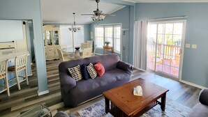 A spacious living room with a dark blue sofa, a wooden coffee table, and a yellow blanket. There is a dining area with a white table and chairs, and a kitchen area with cabinets and a countertop. - Carolina Rose - Steps from the Beach in Oceanside Village (Surfside Beach)