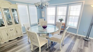 A dining room with a glass table surrounded by four chairs, a white cabinet with glass doors, and a chandelier. - Carolina Rose - Steps from the Beach in Oceanside Village (Surfside Beach)