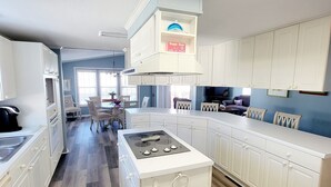 A modern kitchen with white cabinetry, a central island, and a dining area in the background. - Carolina Rose - Steps from the Beach in Oceanside Village (Surfside Beach)