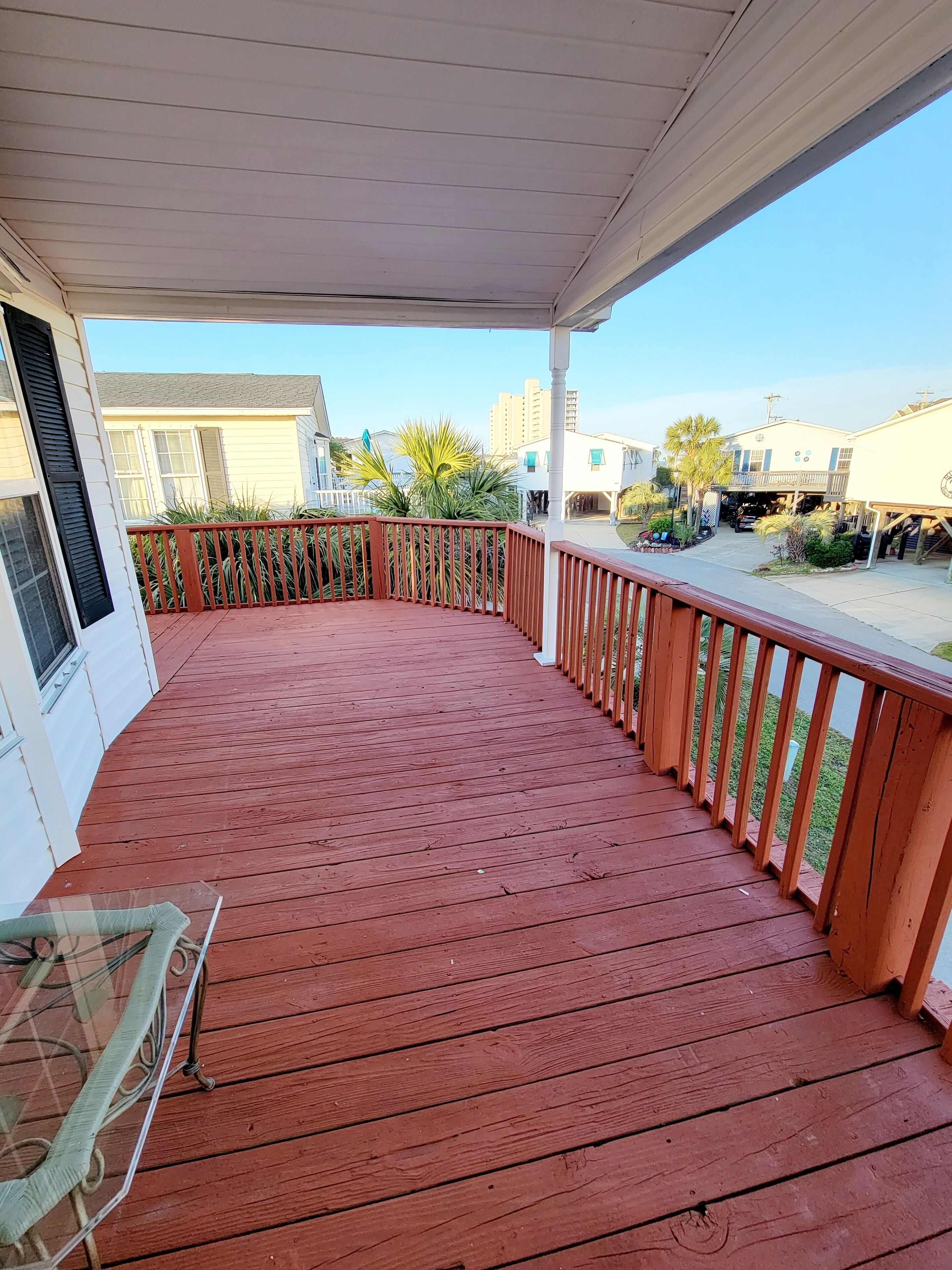 A wooden deck with a railing, a glass table, and a view of a residential area with houses and a clear sky.