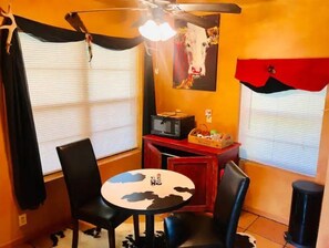 A room with a dining table and chairs, a microwave, a small cabinet, and a painting of a cow on the wall.
