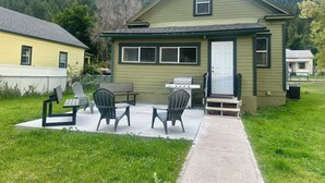 A small, single-story house with a green exterior, a front porch with a table and chairs, and a barbecue grill.