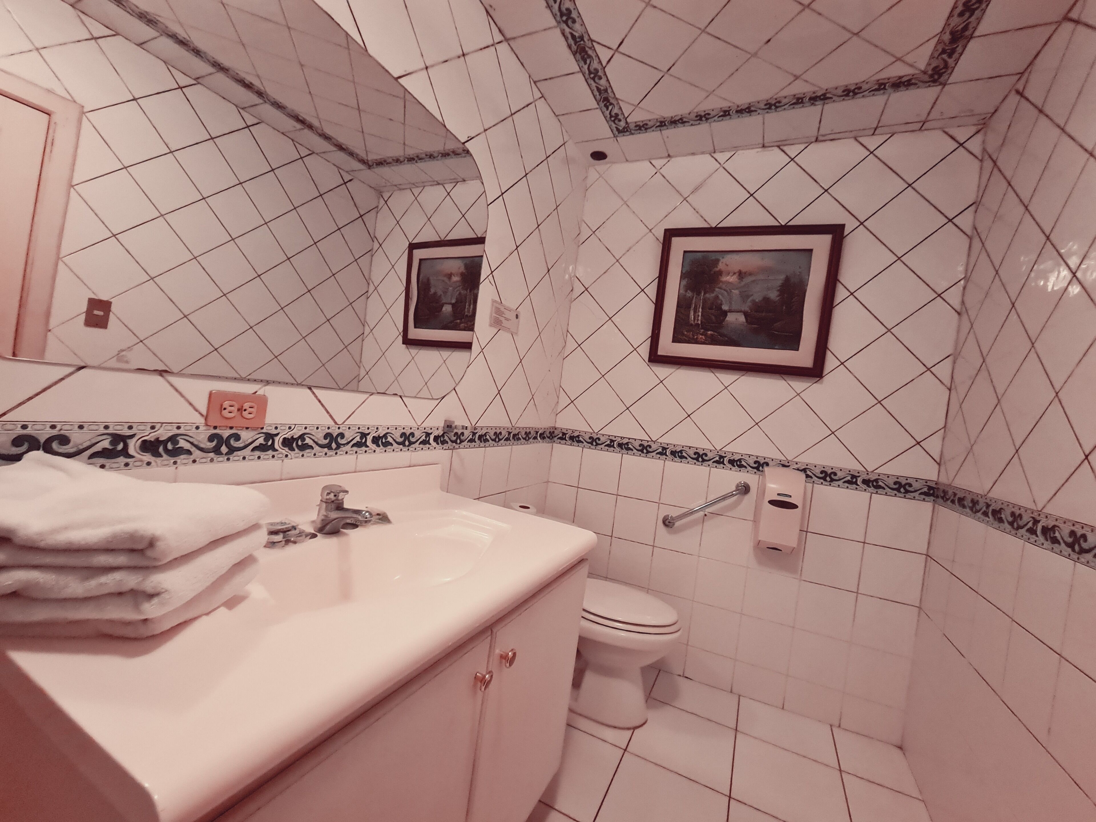 Un baño con paredes de azulejos blancos, un lavamanos con espejo, un inodoro y dos cuadros en la pared.