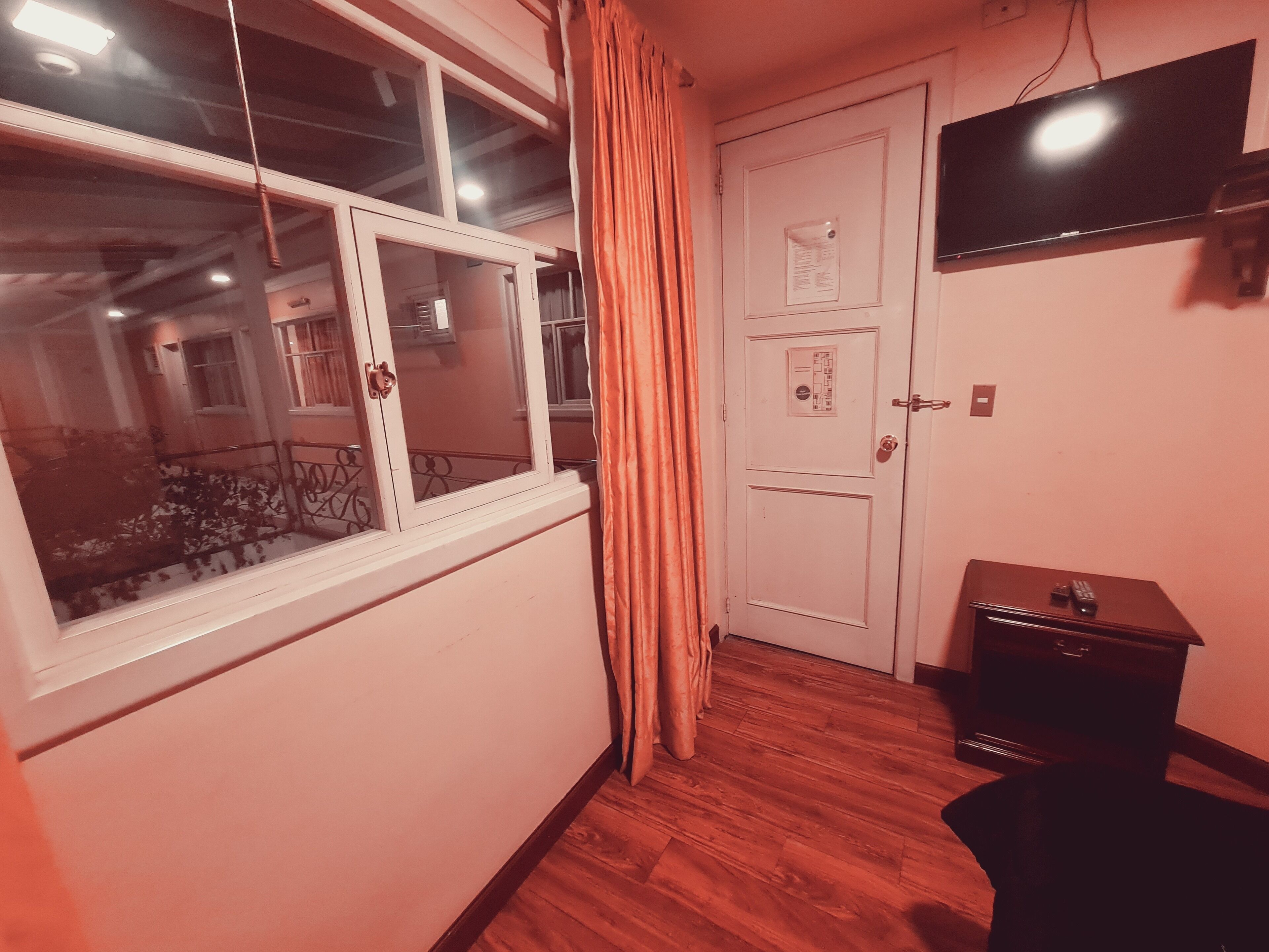 Una habitación con piso de madera, una puerta blanca con cerradura, una ventana con cortina y un televisor de pantalla plana montado en la pared.