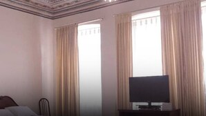 A hotel room with a television on a stand, two windows with curtains, and a bed with a headboard. - Hotel Chat Noir Cuenca (Cuenca)