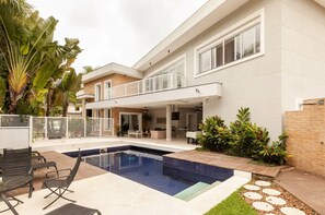 Uma casa moderna de dois andares com piscina, área de estar externa e vegetação exuberante.