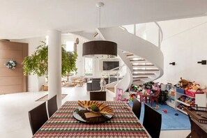 Um interior moderno com uma escada em espiral, uma área de refeições com toalha de mesa estampada e um espaço infantil com brinquedos.