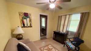 Una sala de estar con un sofá, una silla, una lámpara, un cuadro, un ventilador de techo y una ventana con cortinas.