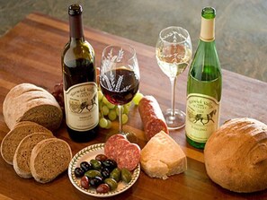 A wooden table with sliced bread, a plate of olives, a bottle of wine, a glass of wine, and a loaf of bread.