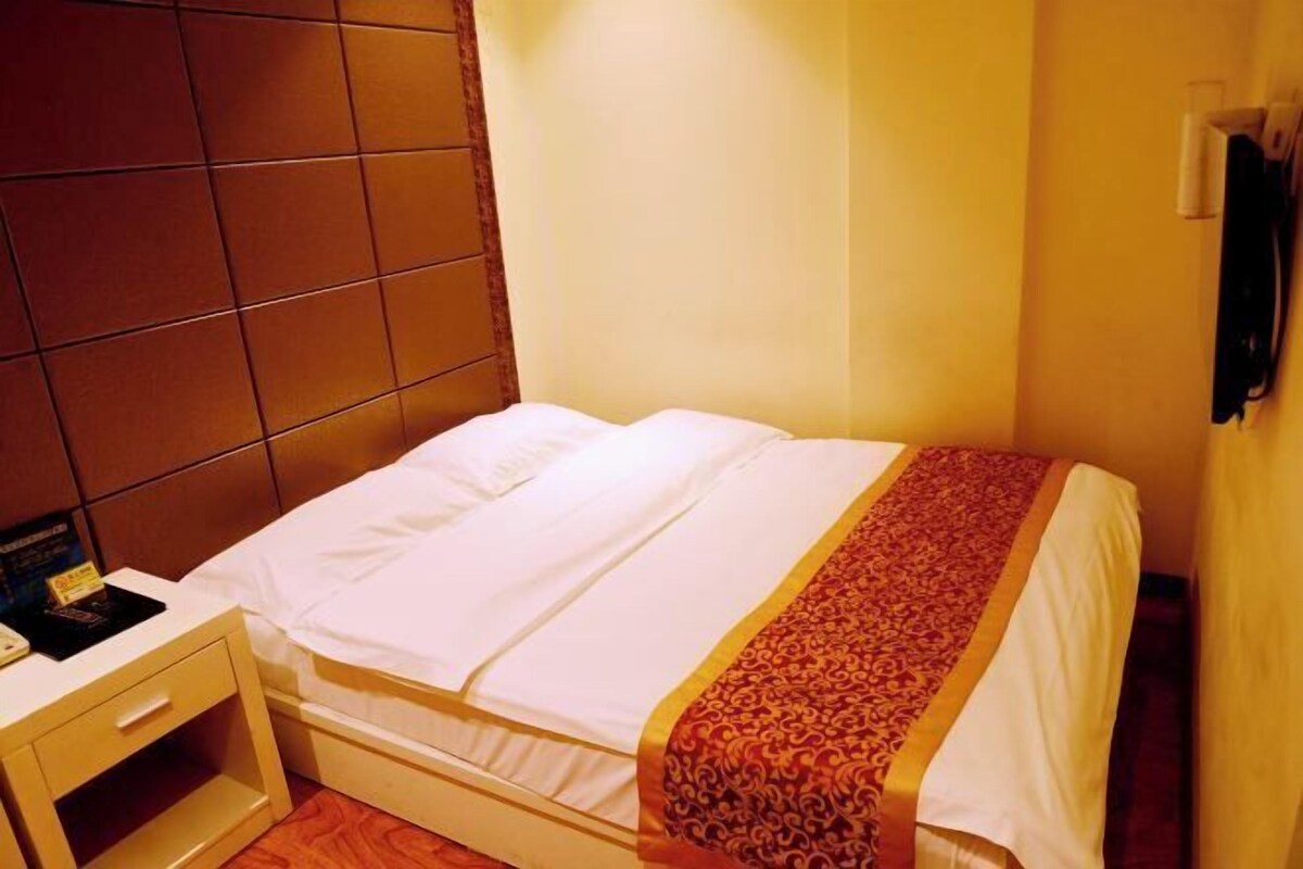 A single bed with a white mattress and a patterned blanket, a bedside table with a lamp, and a wall-mounted towel rack.