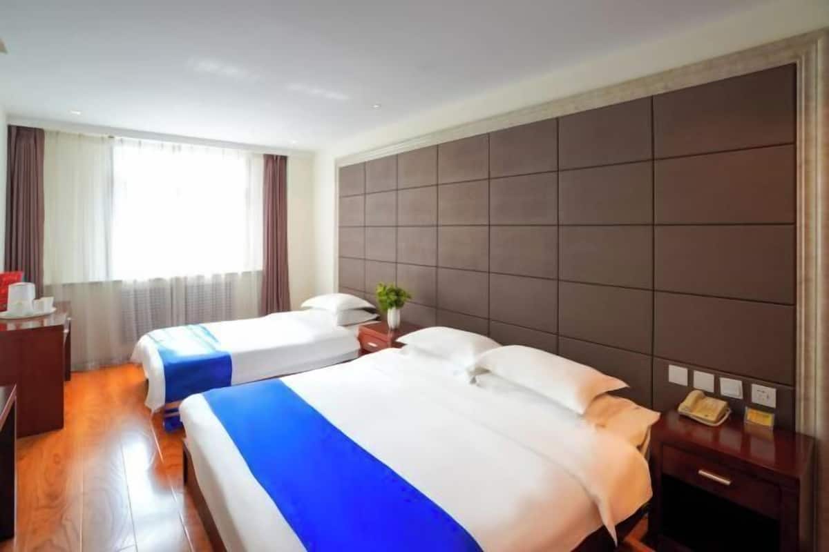 A hotel room with two beds, a wooden floor, a large window with curtains, a bedside table with a lamp, and a wall panel.