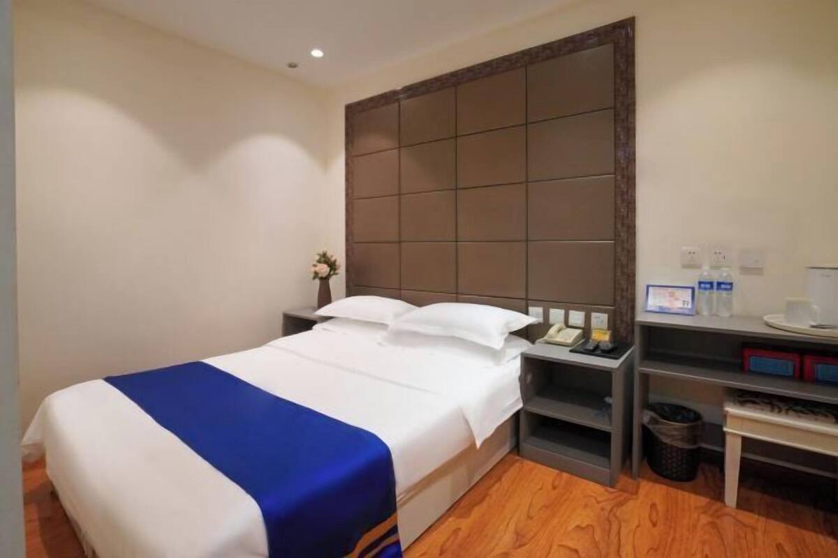 A hotel room with a bed, a nightstand, a shelf with toiletries, and a trash bin.