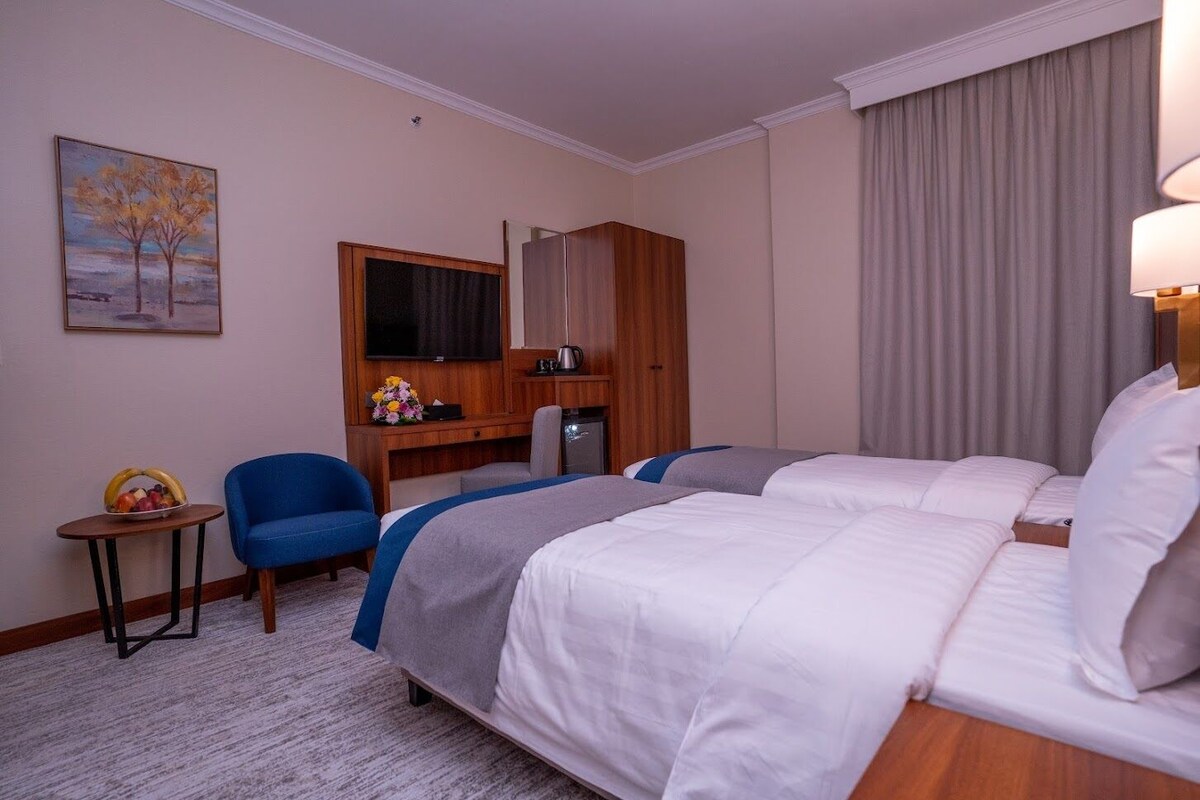 A hotel room with two beds, a desk, a chair, a television, and a painting on the wall.
