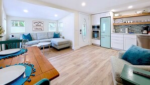A modern kitchen with built-in appliances, a dining area with a wooden table and blue placemats, and a living room with a grey sectional sofa and a decorative glass door.