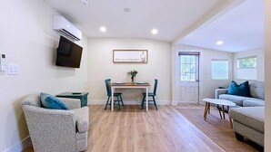 A living room with a dining area, a grey sofa, a patterned armchair, a TV mounted on the wall, and a door with windows.