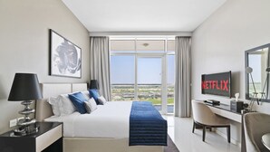 A hotel room with a large bed, a desk, a TV, and a view of the city. - Chic Hotel-Style Studio in Damac Hills (Dubai)
